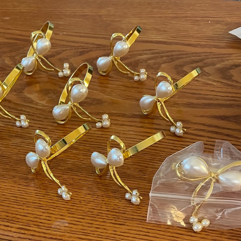 K&ouml;nnte beinhalten: Goldfarbene Metall-Serviettenringe mit Schleifendesign, jeweils mit Kunstperlen verziert. Die Schleife hat eine gro&szlig;e Perlenmitte und kleinere Perlencluster. Mehrere Ringe sind sichtbar, einige in einer durchsichtigen Plastikt&uuml;te.