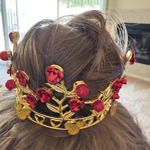 Könnte beinhalten: Eine goldfarbene Krone mit roten Rosen und Edelsteinakzenten. Die Krone hat ein Herz- und Mickey-Mouse-Design. Die Krone wird auf braunem Haar getragen.