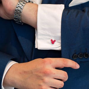 May include: A man wearing a dark blue suit jacket with a white dress shirt and a silver watch. The shirt has pink Mickey Mouse cufflinks.