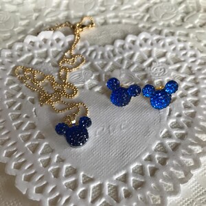 May include: A gold-colored necklace and matching earrings set. The pendant and earrings are shaped like a mouse head, encrusted with dark blue crystals. The jewelry is displayed on a white, lacy surface.