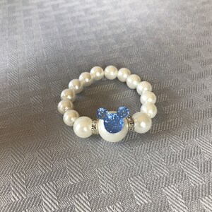Puede incluir: Una pulsera de perlas blancas con un dije azul en forma de Mickey Mouse. El dije est&aacute; adornado con pedrer&iacute;a azul y rodeado por una base blanca. La pulsera est&aacute; hecha de perlas blancas redondas.