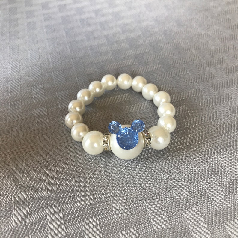Puede incluir: Una pulsera de perlas blancas con un dije azul en forma de Mickey Mouse. El dije est&aacute; adornado con pedrer&iacute;a azul y rodeado por una base blanca. La pulsera est&aacute; hecha de perlas blancas redondas.
