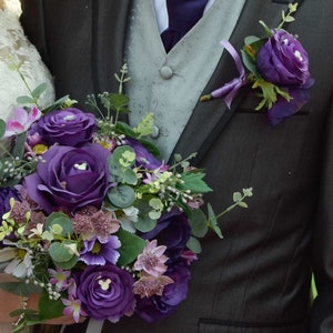 May include: A bridal bouquet and boutonniere featuring deep purple roses and other flowers. The bouquet is held by a person wearing a white lace dress. The boutonniere is pinned to a dark gray suit jacket.
