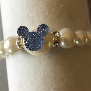 Puede incluir: Una pulsera de perlas blancas con un charm de Mickey Mouse de cristal azul. La pulsera presenta perlas grandes y redondas y detalles plateados. El charm es de color azul claro y est&aacute; hecho de peque&ntilde;os cristales.
