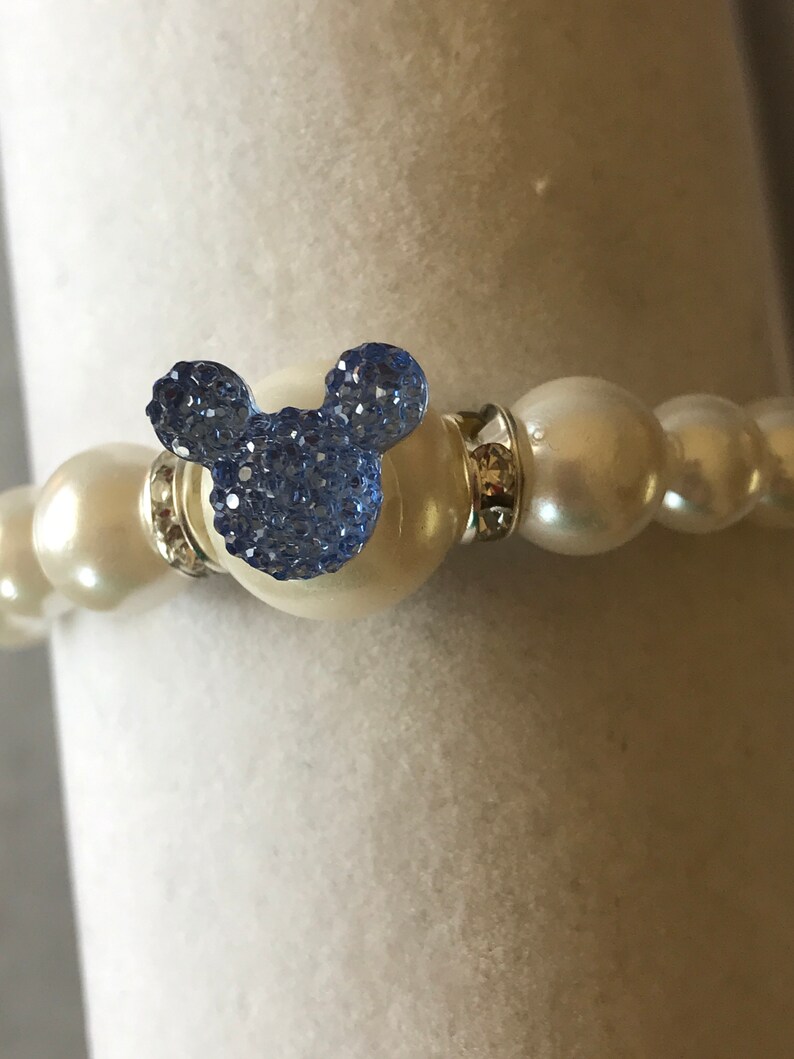 Puede incluir: Una pulsera de perlas blancas con un charm de Mickey Mouse de cristal azul. La pulsera presenta perlas grandes y redondas y detalles plateados. El charm es de color azul claro y est&aacute; hecho de peque&ntilde;os cristales.