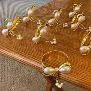 K&ouml;nnte beinhalten: Eine Sammlung goldfarbener Serviettenringe mit Perlen- und Schleifendesign. Die Serviettenringe sind auf einer Holzoberfl&auml;che angeordnet und pr&auml;sentieren ihren eleganten und dekorativen Stil. Die Perlen sind wei&szlig; und das Metall hat ein gl&auml;nzendes Finish.