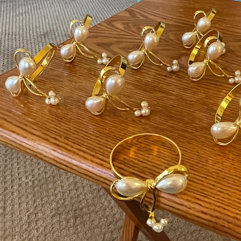 K&ouml;nnte beinhalten: Eine Sammlung goldfarbener Serviettenringe mit Perlen- und Schleifendesign. Die Serviettenringe sind auf einer Holzoberfl&auml;che angeordnet und pr&auml;sentieren ihren eleganten und dekorativen Stil. Die Perlen sind wei&szlig; und das Metall hat ein gl&auml;nzendes Finish.
