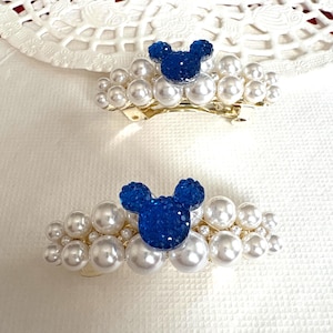 May include: Two hair clips with a blue, sparkly mouse head design on a bed of faux pearls. The clips have a gold-coloured metal base. The background is a white surface with a lace doily.