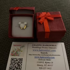 May include: A small, white, sparkly Mickey Mouse head hair clip in a red gift box. The box has a red ribbon and bow. The hair clip is on a white background. The text on the card reads: "AMAZING HAIRSWIRLS Weddings Proms Dancers www.hairswirls1.com Search: http://stores.ebay.com/hairjewelrydebstwisties www.hairswirls.com Deborah Miller 13109 E. Ramos St Dewey, AZ 86327 928-227-3612"