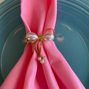K&ouml;nnte beinhalten: Ein goldfarbener Serviettenring in Schleifenform, verziert mit zwei gro&szlig;en perlmuttartigen Perlen und kleineren Perlen. Der Serviettenring h&auml;lt eine rosa Serviette, die auf einem hellblauen Teller liegt. Dieses elegante Accessoire verleiht jedem Tisch einen Hauch von Eleganz.