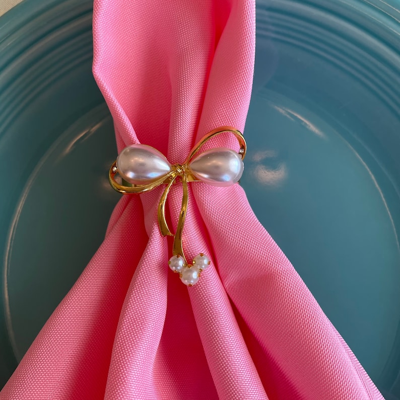 K&ouml;nnte beinhalten: Ein goldfarbener Serviettenring in Schleifenform, verziert mit zwei gro&szlig;en perlmuttartigen Perlen und kleineren Perlen. Der Serviettenring h&auml;lt eine rosa Serviette, die auf einem hellblauen Teller liegt. Dieses elegante Accessoire verleiht jedem Tisch einen Hauch von Eleganz.