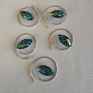 Könnte beinhalten: Vier silberfarbene Weinanhänger, jeweils mit einem blattförmigen, irisierenden blau-grünen Edelstein. Die Edelsteine sind in einem silbernen Spiraldesign gefasst, mit einem kleinen, durchsichtigen Kunststoffteil am Ende.