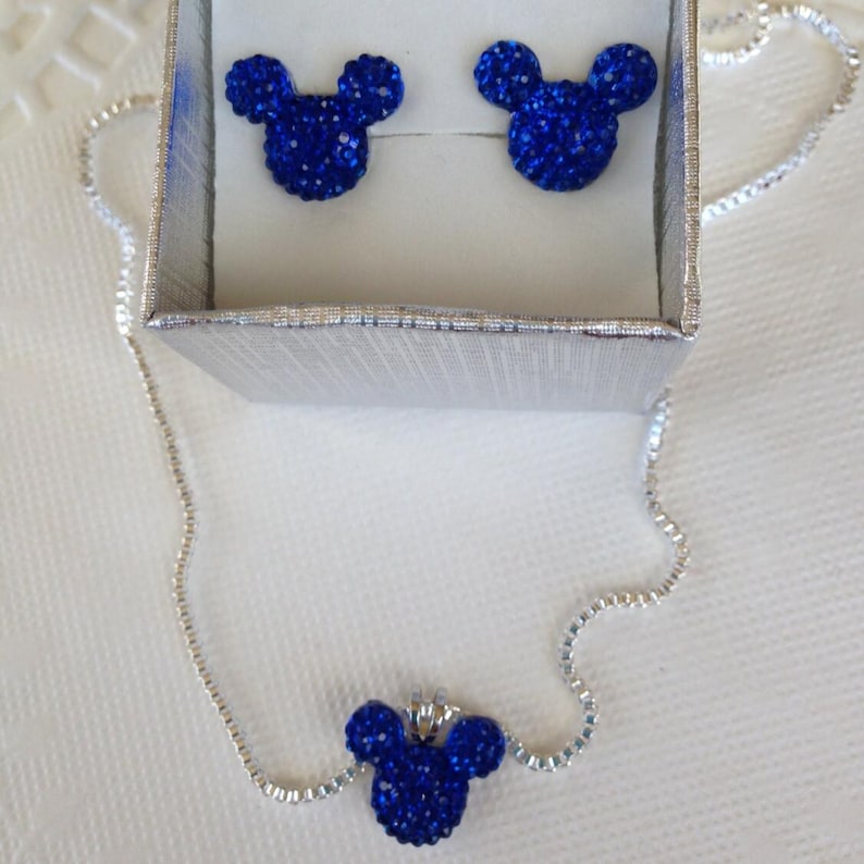 A silver necklace and earrings set with blue crystal Mickey Mouse charms. The necklace is a silver chain with a blue crystal Mickey Mouse charm pendant. The earrings are silver posts with blue crystal Mickey Mouse charms.