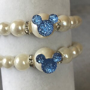 Puede incluir: Dos pulseras de perlas con dijes de cabeza de Mickey Mouse de cristal azul. Las pulseras tienen perlas grandes, redondas, blanquecinas y espaciadores plateados con cristales transparentes. Los dijes son de color azul claro.