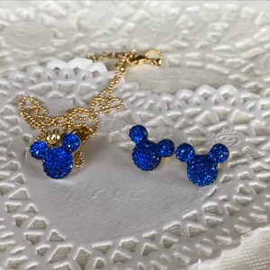 May include: A gold-colored necklace and matching earrings set. The pendant and earrings are shaped like a mouse head, encrusted with sparkling blue crystals. The jewelry set is displayed on a white, lacy surface.
