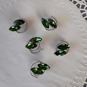 Könnte beinhalten: Fünf grüne Kristall-Haarspiralen. Jede Spirale hat zwei tropfenförmige grüne Kristalle auf einem silberfarbenen Metallsockel. Die Kristalle haben eine facettierte Oberfläche, die das Licht reflektiert. Die Haaraccessoires sind auf einer weißen, strukturierten Oberfläche angeordnet.