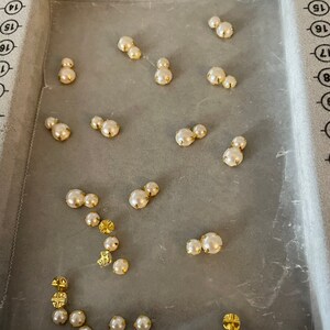 K&ouml;nnte beinhalten: Eine Sammlung kleiner, cremefarbener Perlen mit goldfarbenen Metalleinfassungen. Die Perlen sind auf einer hellgrauen Oberfl&auml;che angeordnet. Einige Perlen sind einzeln, andere paarweise oder in Gruppen angeordnet. Die goldenen Fassungen verleihen Eleganz.