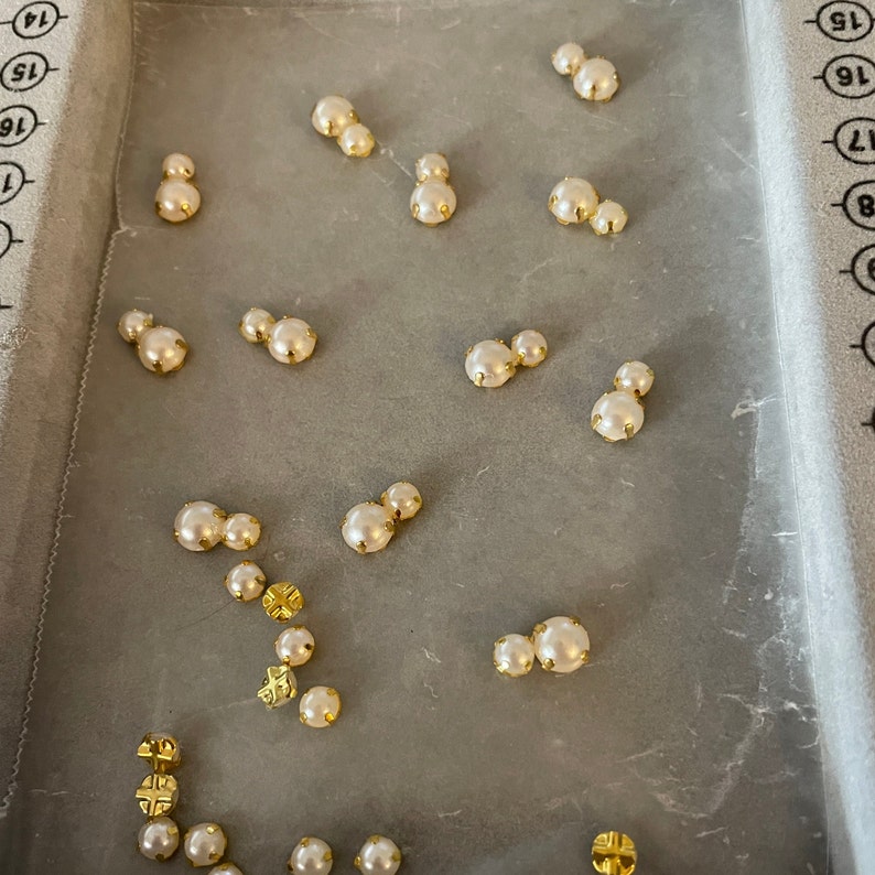 K&ouml;nnte beinhalten: Eine Sammlung kleiner, cremefarbener Perlen mit goldfarbenen Metalleinfassungen. Die Perlen sind auf einer hellgrauen Oberfl&auml;che angeordnet. Einige Perlen sind einzeln, andere paarweise oder in Gruppen angeordnet. Die goldenen Fassungen verleihen Eleganz.
