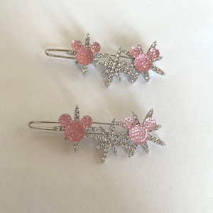 May include: Two silver hair clips, each featuring a pink, glittery mouse head design and a sparkling starburst accent. The clips are set against a white background, showcasing the decorative hair accessories.