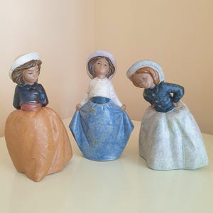 May include: Three ceramic figurines of individuals wearing hats and long skirts. The left figure wears a dark top and orange skirt, the center figure wears a white top and blue skirt, and the right figure wears a dark top and light patterned skirt.