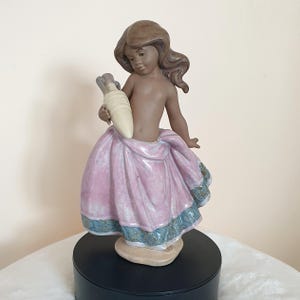 May include: A ceramic figurine of a young girl holding a cream-colored object, possibly a vase or a horn. She is wearing a pink skirt with blue and green trim. The figurine has brown hair and skin, and is standing on a black base.