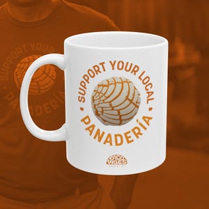 May include: White ceramic mug with a brown graphic that says "Support Your Local Panaderia" with a concha bread illustration. The text "Taco Vibes Apparel" is printed in brown at the bottom of the mug.