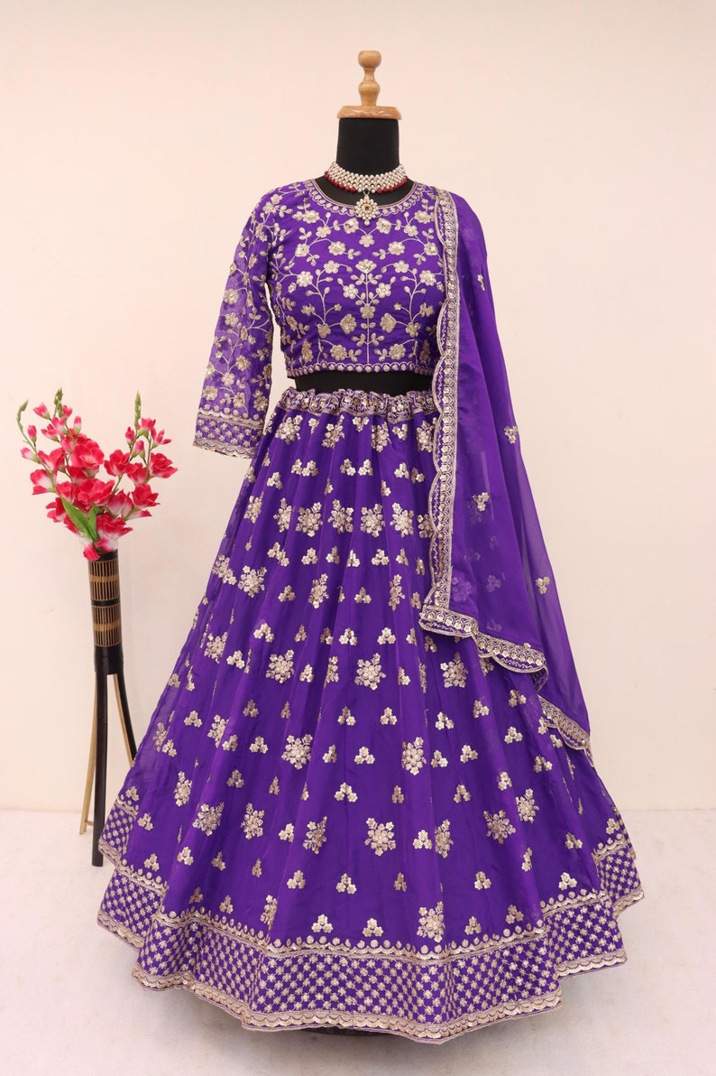 May include: A vibrant purple lehenga, a traditional Indian garment, displayed on a mannequin. The ensemble features intricate gold embroidery on the blouse, skirt, and dupatta. The skirt has a full, flowing design, and the dupatta is draped over the shoulder.