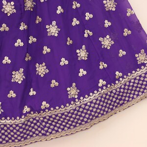 May include: Close-up of a vibrant purple fabric with intricate gold embroidery. The design features floral patterns and a detailed border, creating a luxurious and ornate appearance. This textile is suitable for formal wear or decorative projects.