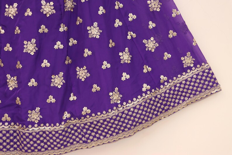 May include: Close-up of a vibrant purple fabric with intricate gold embroidery. The design features floral patterns and a detailed border, creating a luxurious and ornate appearance. This textile is suitable for formal wear or decorative projects.