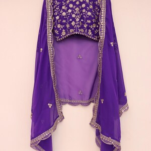 May include: A purple ensemble featuring a heavily embroidered top with gold floral patterns. The outfit includes a matching sheer purple shawl with gold trim and embellishments. The clothing is displayed on a wooden hanger against a plain white background.