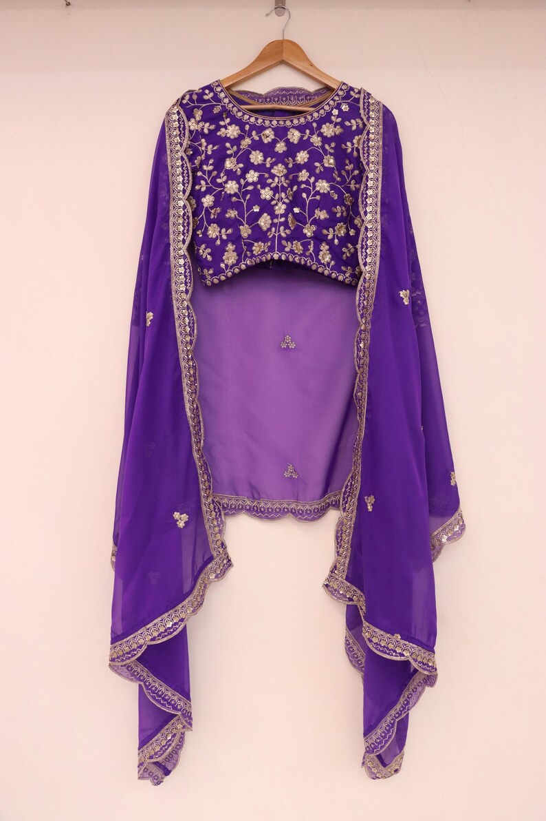 May include: A purple ensemble featuring a heavily embroidered top with gold floral patterns. The outfit includes a matching sheer purple shawl with gold trim and embellishments. The clothing is displayed on a wooden hanger against a plain white background.