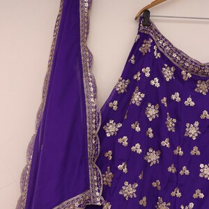 May include: A vibrant purple ensemble featuring a skirt and a matching scarf. The skirt is adorned with intricate gold floral embroidery. The scarf has a scalloped edge with gold trim. The outfit is hanging on a wooden hanger.