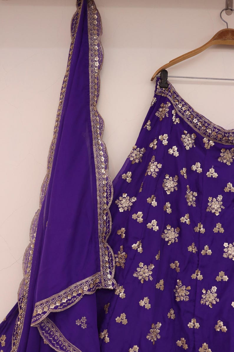 May include: A vibrant purple ensemble featuring a skirt and a matching scarf. The skirt is adorned with intricate gold floral embroidery. The scarf has a scalloped edge with gold trim. The outfit is hanging on a wooden hanger.