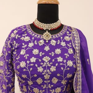 May include: A purple embroidered blouse and skirt set with gold floral patterns. The outfit is displayed on a mannequin and includes a matching purple scarf. A pearl and red bead necklace with a pendant is also featured.