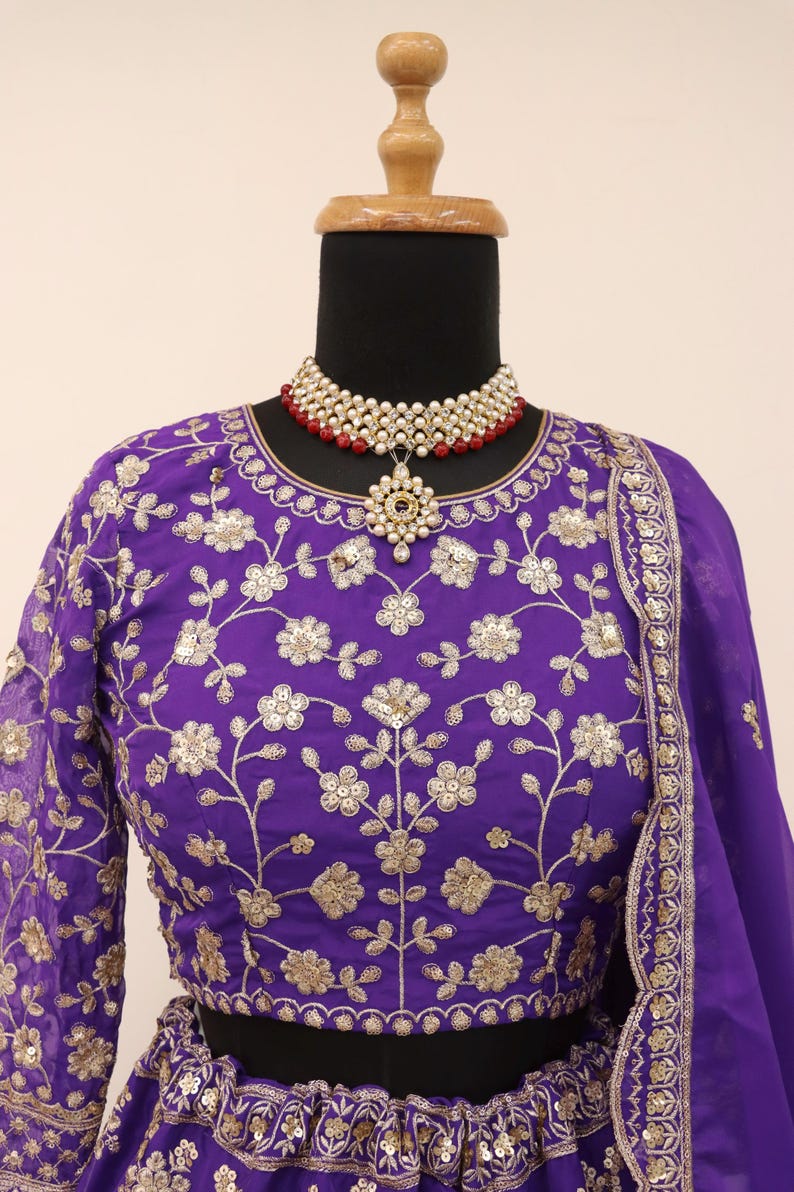 May include: A purple embroidered blouse and skirt set with gold floral patterns. The outfit is displayed on a mannequin and includes a matching purple scarf. A pearl and red bead necklace with a pendant is also featured.