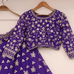 May include: A purple embroidered outfit, including a long-sleeved top and a skirt, hangs on wooden hangers. The outfit features intricate floral embroidery in a contrasting color, creating a detailed and elegant design. The fabric appears to be a rich, luxurious material.