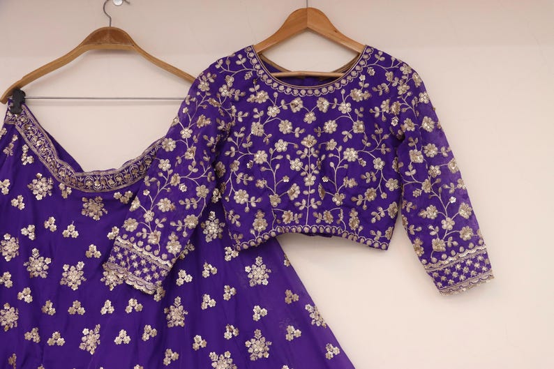 May include: A purple embroidered outfit, including a long-sleeved top and a skirt, hangs on wooden hangers. The outfit features intricate floral embroidery in a contrasting color, creating a detailed and elegant design. The fabric appears to be a rich, luxurious material.