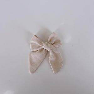 May include: A beige fabric bow, likely for hair, is centered against a white background. The bow is tied in the middle, with two loops and two long, pointed ends. The fabric has a textured appearance.