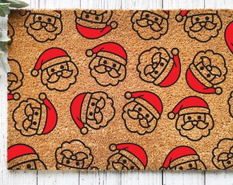 May include: A brown coir doormat with a repeating pattern of red Santa Claus faces. The Santa faces are outlined in black and have white eyes and a white beard.