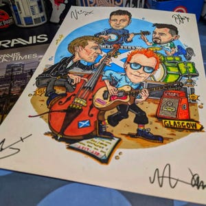 Puede incluir: Una ilustración dibujada a mano de una banda tocando música. Los miembros de la banda están dibujados en un estilo de dibujos animados y visten ropa colorida. La ilustración está ambientada en un fondo de un escenario con un micrófono y una batería. El nombre de la banda, "TRAVIS", está escrito en letras grandes en la parte superior de la ilustración.