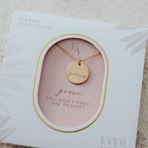 May include: A 14 karat gold-filled necklace with a round pendant engraved with the word grace. The jewelry is presented in a white square box with a gold-rimmed oval cutout, displaying the text grace and YOU DON'T HAVE TO BE PERFECT.