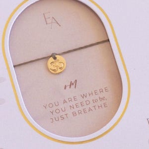 Puede incluir: Una tarjeta blanca con letras doradas y un brazalete con un dije dorado. La tarjeta dice "14 Karat Gold-Filled", "EA", "You are where you need to be, Just Breathe", "Ever Aster". El dije es un símbolo Om dorado.