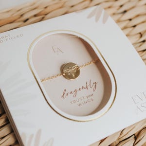 Könnte beinhalten: Ein goldfarbenes Armband mit einem Libellen-Anhänger und den Worten "dragonfly trust your wings" auf einer weißen Karte mit goldfarbenem Rand.