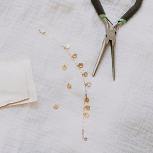 Puede incluir: Un collar de cadena dorada con pequeños colgantes redondos. El collar está sobre una superficie de tela blanca con unas pinzas de plata al lado.