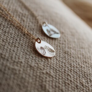 Puede incluir: Dos collares de oro con cadenas delicadas y colgantes ovalados con un dise&ntilde;o de hoja detallado. Los collares se muestran sobre un fondo de tela beige.