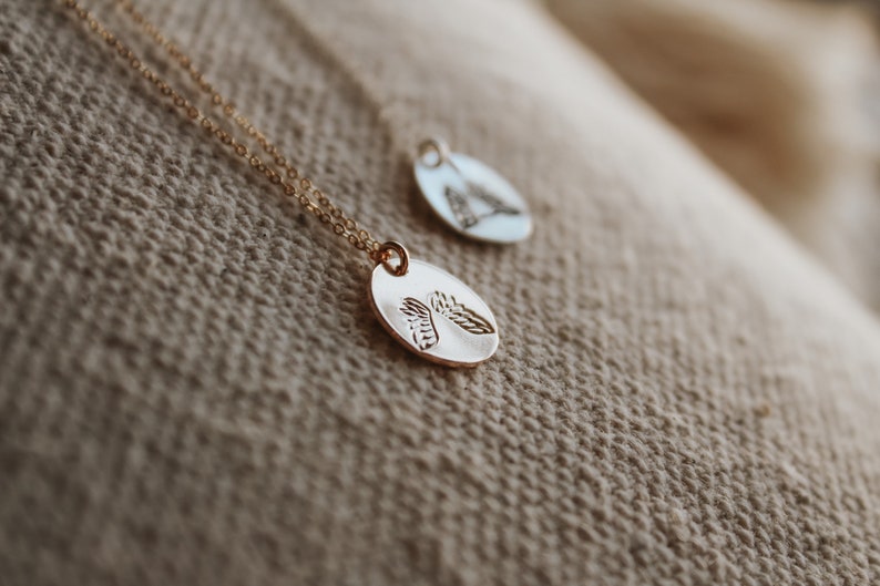 Puede incluir: Dos collares de oro con cadenas delicadas y colgantes ovalados con un dise&ntilde;o de hoja detallado. Los collares se muestran sobre un fondo de tela beige.