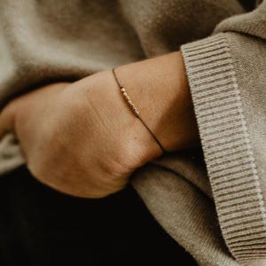 May include: A simple bracelet with a delicate chain of small gold beads on a black cord.