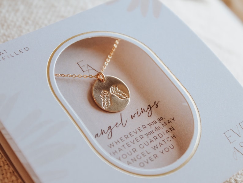 Puede incluir: Un collar de oro con un colgante redondo que presenta un par de alas de &aacute;ngel. El collar se muestra en una caja blanca con el texto "angel wings" y un mensaje sobre un &aacute;ngel guardi&aacute;n que te cuida.
