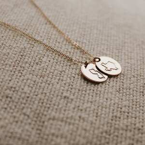 May include: A gold-coloured necklace with two oval pendants. Each pendant has a state outline engraved on it. The chain and pendants have a shiny finish. The necklace is displayed on a textured, neutral-coloured surface.