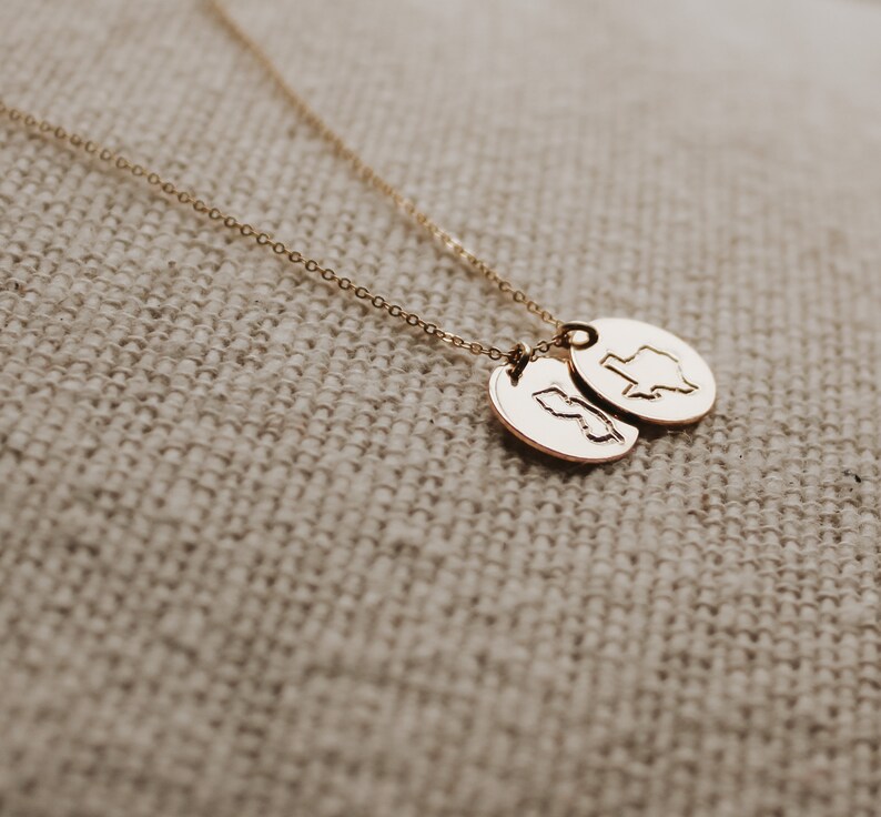 May include: A gold-coloured necklace with two oval pendants. Each pendant has a state outline engraved on it. The chain and pendants have a shiny finish. The necklace is displayed on a textured, neutral-coloured surface.